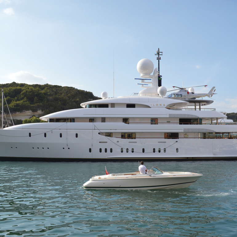 Large white superyacht moored off a wooded coast with a tender passing by.