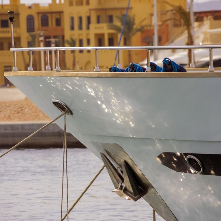 Bow of a white yacht reflected in calm water in front of waterfront buildings.