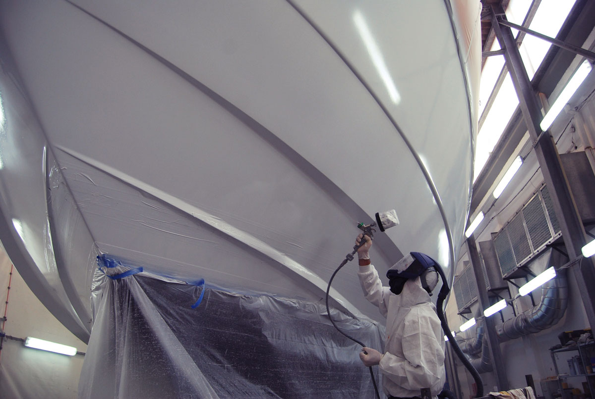 Painter in full protective suit spraying the hull of a yacht in a shipyard.