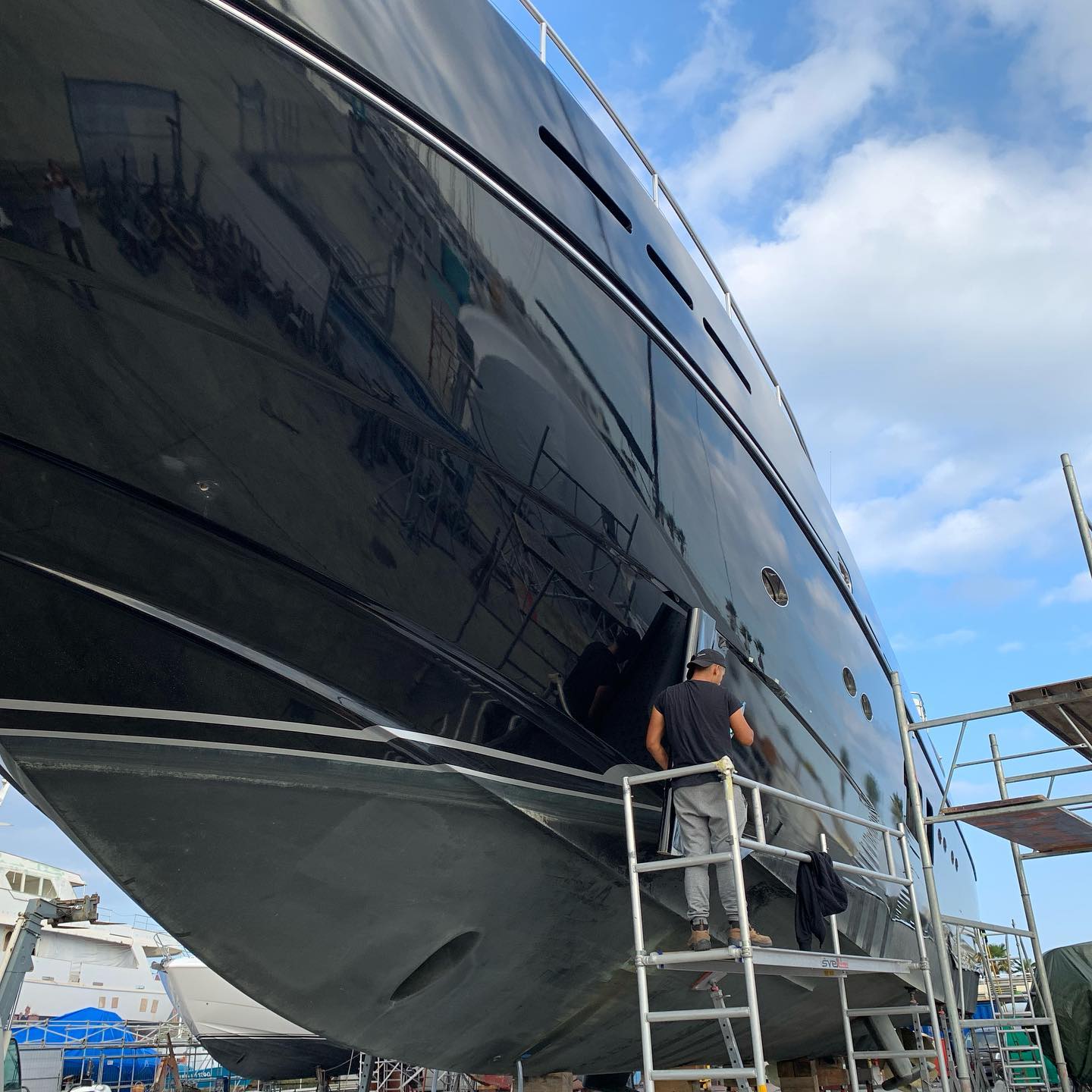 Technician on scaffolding working on a mirror-finish black yacht hull.