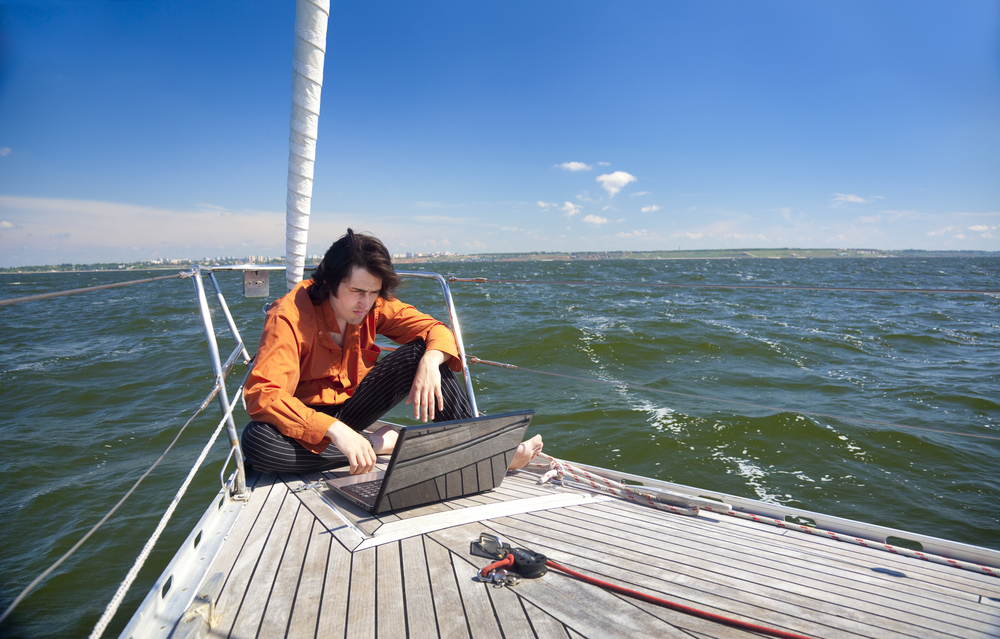 Man working on a laptop in the cockpit of a sailing yacht at sea.