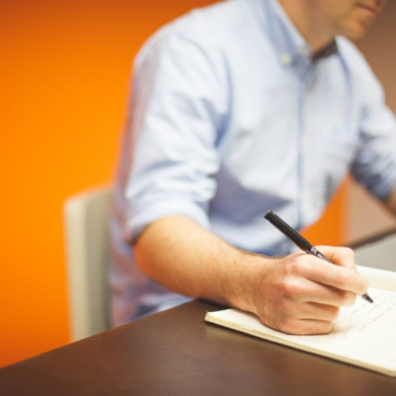 Person writing notes in a notebook beside a laptop.